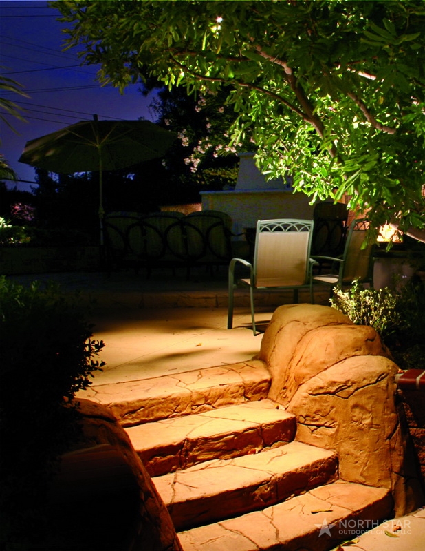 Flagstone patio steps lit from the side by low-voltage fixtures, with patio seating and an uplit tree canopy beyond