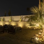 Lit backyard patio seating with uplit palms and a washed block wall at night