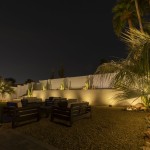 Backyard patio lit at night with uplit palms and cactus against a block wall