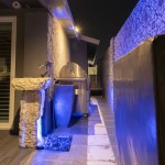 Side yard with a barbecue and trash bins lit in blue accent lighting at night