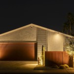 Ranch home garage elevation lit at night with a rust-metal fence and uplit cactus