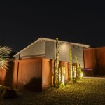 Rust-metal fence and uplit columnar cactus in front of a lit home at night