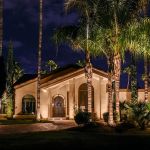 Stucco home with an arched entry lit at night beneath uplit palms