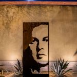 Backlit metal portrait panel mounted on a wall, lit at night with agave below