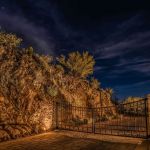 Iron driveway gate and stone wall lit at night beside an uplit rock face