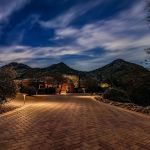 Paver drive lit at night leading to an adobe home with mountains behind