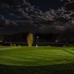 Golf green with a flagstick lit at night beneath a cloudy sky