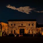 Two-story stone home with a lit entry and uplit desert planting at night