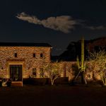 Stone home lit at night with an uplit saguaro cactus beside it
