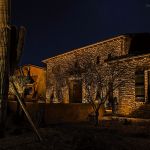 Stone and stucco home lit at night with uplit cactus and trees along the entry