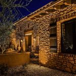 Stone-faced home entrance lit at night with wall sconces and uplit planting