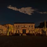 Stone estate home lit at night with uplit cactus and a cloud overhead