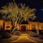 Uplit mesquite tree over a flagstone path leading to a lit adobe home entrance