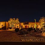 Long adobe home lit at night with uplit trees and cactus along the front