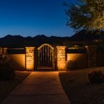 Iron pedestrian gate in a stone wall lit at night above a paver walkway