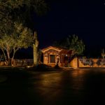 Community entrance building and gate lit at night with uplit saguaro and trees
