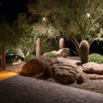 Uplit boulders, barrel cactus and desert trees beside a paver walkway at night