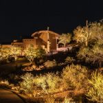 Hilltop desert home lit at night with uplit trees and cactus on the slope below