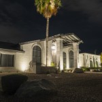 White stucco home lit at night with an uplit palm and lawn in the foreground