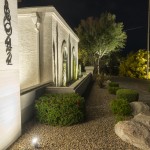 Side elevation of a lit white stucco home at night with uplit shrubs and boulders