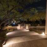 Lit paver driveway and courtyard gate at night with uplit trees