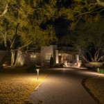 Path lights and uplit trees along a driveway to a lit home at night
