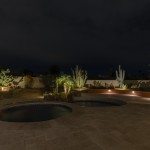 Lit pool and spa in a backyard at night with uplit desert planting