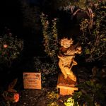 Lit garden statue of a cherub with a lyre beside a memorial plaque at night